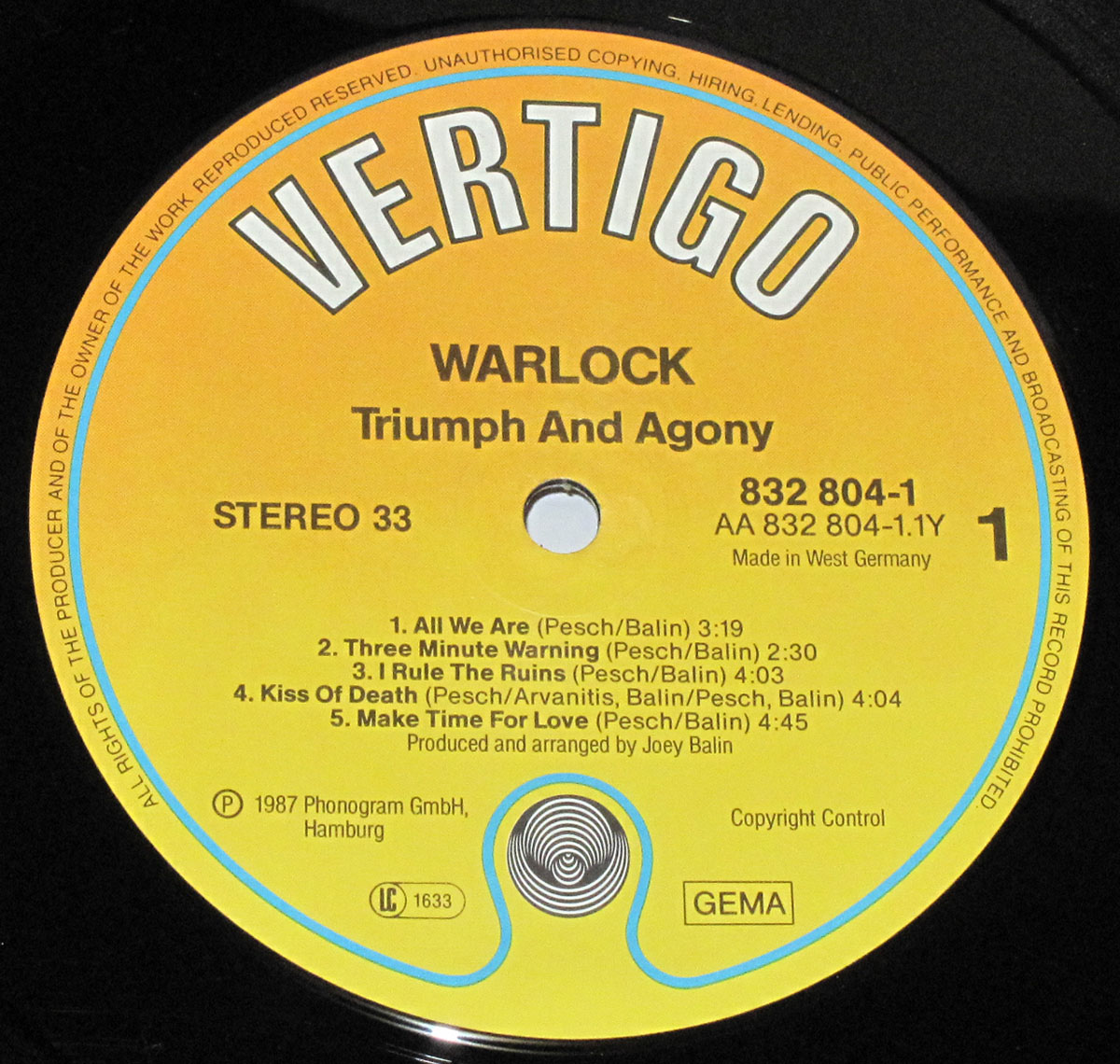 Side One record label of Warlock’s 1987 album Triumph and Agony, West German Vertigo pressing. The label is a bright yellow-to-orange gradient with bold Vertigo logo across the top. Below, the album title and band name are printed in black, followed by catalog number 832 804-1 / AA 832 804-1.1Y, stereo speed designation, and track list for Side One: All We Are, Three Minute Warning, I Rule the Ruins, Kiss of Death, and Make Time for Love. The lower section displays the 1987 Phonogram GmbH copyright, LC1633 code, GEMA rights society mark, and the distinctive black-and-white Vertigo swirl logo at 6 o’clock.
